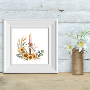 Op de afbeelding: Een ingelijste aquarelprint met een kaars in een gouden kandelaar, omringd door zonnebloemen, tarwe en groen. Een label met de tekst "Blessed" hangt aan de kandelaar. De print zit in een witte lijst, met rechts een vaas met witte bloemen.