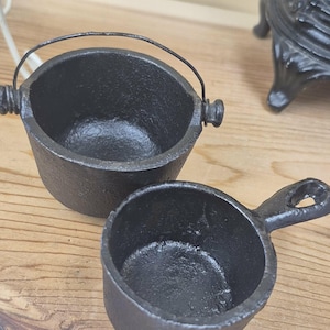 May include: Two small black cast iron cooking vessels. One is a round pot with a thin wire handle attached to two side lugs. The other is a small round pan featuring a short handle with a hole at the end for hanging. Both have a textured finish.