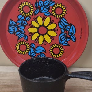 May include: A round red metal tray featuring a central yellow flower surrounded by smaller yellow flowers and blue leaves, with the text Made in Brazil printed on the side. In front sits a small black cast iron pot with a single handle.