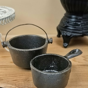 May include: A miniature black cast iron cauldron with a thin metal wire handle and a small black cast iron skillet with a short handle. Both items feature a textured, dark metallic finish and are designed as small-scale kitchenware replicas.