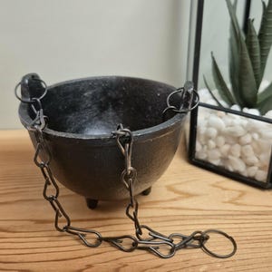 May include: A black cast iron cauldron with three small feet and two side handles. A dark metal chain is attached to the handles, draping down in front of the bowl. The cauldron has a smooth, rounded interior and a textured exterior surface.