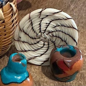 May include: A woven basket, a circular woven coaster with dark brown accents, and two small, hand-painted ceramic candle holders. The candle holders are brown, orange, and turquoise. The basket is made of light brown woven material.