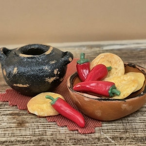 May include: A miniature black pot with two handles sits beside a small brown bowl filled with several bright red chili peppers and round, pale yellow flatbreads. One additional red chili pepper rests on a textured red fabric piece.