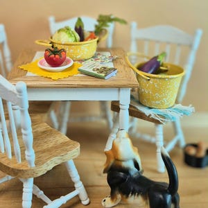 May include: Miniature kitchen scene with a small wooden table and white chairs. A yellow pot and bucket hold vegetables. A ceramic beagle figurine stands near the table. A small book and a tomato on a plate are also on the table.