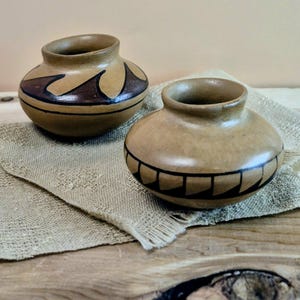 May include: Two small, round, tan-colored pottery vases. One vase has a dark brown and black wave design, and the other has a black geometric pattern around the rim. They sit on a textured, beige cloth.