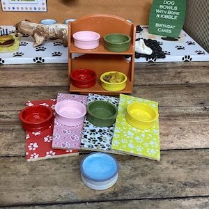 May include: A miniature two-tiered orange shelf holding pink, green, red, and yellow bowls. Below, four rectangular mats with paw print patterns in red, pink, green, and yellow sit alongside a single light blue bowl on a wooden surface.