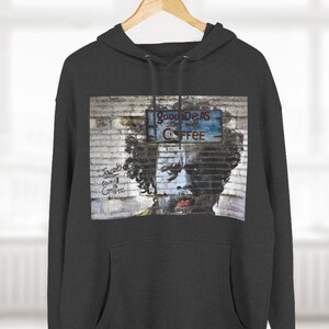 Peut inclure: Un sweat à capuche gris foncé avec un imprimé graphique d'un mur de briques avec une pancarte qui dit "Good Ideas start with Coffee" et une personne avec une barbe et des cheveux bouclés. La personne regarde la caméra.