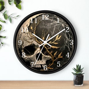 Puede incluir: Reloj de pared negro con un diseño de calavera blanca y ramas doradas. La esfera del reloj tiene números y manecillas blancas.