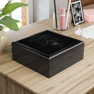 May include: A black wooden box with a glossy finish. The lid features a detailed, embossed design of a skull adorned with roses. The box is rectangular and sits on a wooden surface.