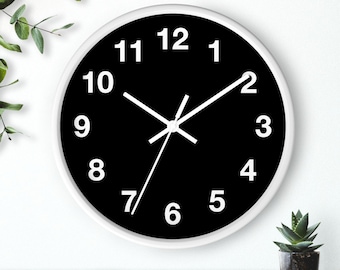 May include: A round wall clock with a white frame and a black face. The clock features white numerals and hands. The clock is mounted on a white wall, with a small potted plant visible in the lower right corner.