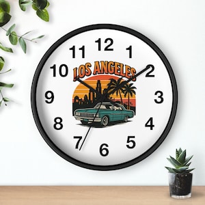 May include: Round wall clock with a black frame and a white face. The clock has black numerals and the words "LOS ANGELES" in orange. A teal car is in front of a sunset with palm trees and a city skyline.