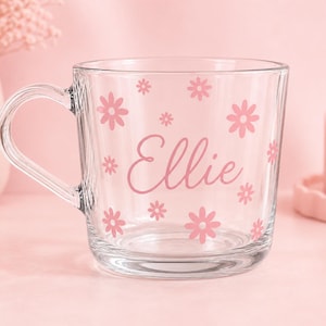 May include: A clear glass mug featuring a pink floral pattern and the name Ellie printed in a cursive script. The mug has a rounded handle and a smooth, transparent finish, with the pink flowers scattered across the front surface of the glass.