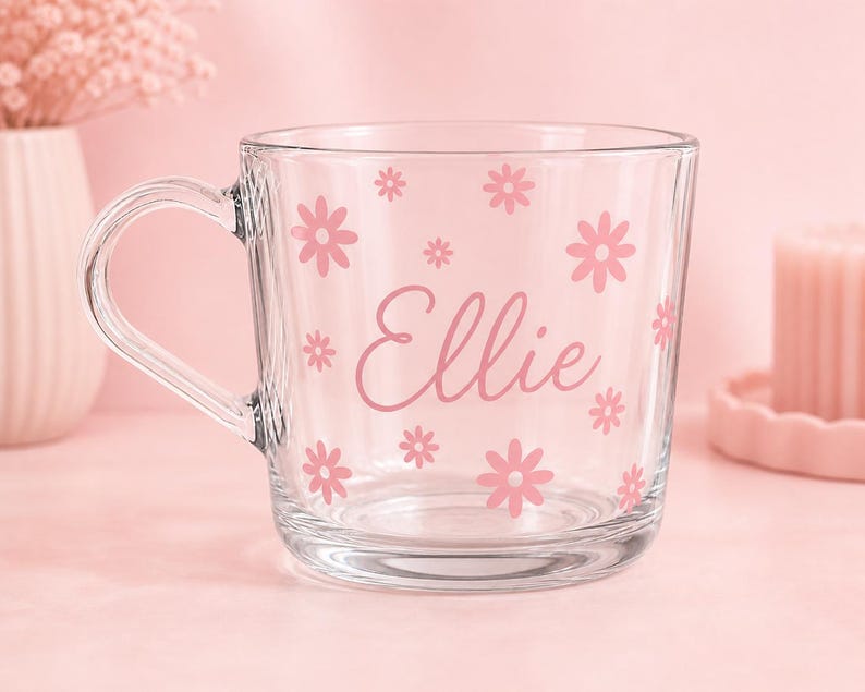 May include: A clear glass mug featuring a pink floral pattern and the name Ellie printed in a cursive script. The mug has a rounded handle and a smooth, transparent finish, with the pink flowers scattered across the front surface of the glass.
