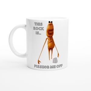 May include: White ceramic mug with a cartoon character and a small rock. The text on the mug reads "THIS ROCK IS... PISSING ME OFF." The character is brown with long arms and legs.