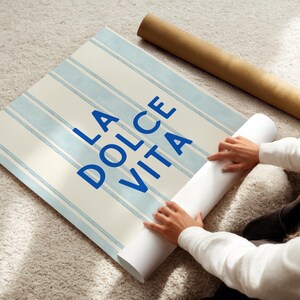 Può includere: Un rotolo di carta da parati a righe blu e bianche con la scritta "LA DOLCE VITA" in blu. Un secondo rotolo di carta da parati è visibile sullo sfondo. La carta da parati viene srotolata su un tappeto di colore chiaro.