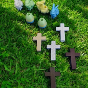 May include: A selection of Easter decorations on green grass. The image shows colourful bunny figurines, egg-shaped candle holders, and crosses in various colours, including white, brown, and tan. The scene is lit by natural sunlight.
