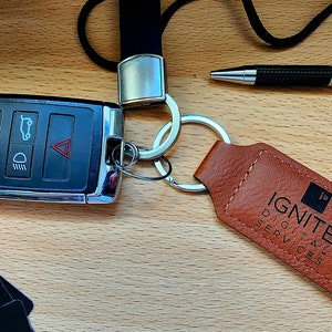 Puede incluir: Un llavero de cuero marrón con el texto "IGNITE DIGITAL SERVICES" y un logotipo negro. El llavero está unido a un llavero plateado y a un cordón negro.