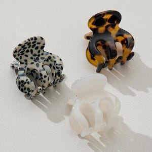 May include: Three claw hair clips in the image. One clip has a black and white spotted pattern, another has a tortoiseshell design, and the third is solid white. These small clips are designed to hold hair in place.