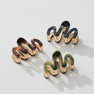 May include: Three wavy hair claws featuring a gold-tone metal base and a patterned resin top. One claw has a brown tortoiseshell pattern, one is solid black with subtle grey marbling, and one is a deep olive green with dark marbled accents.