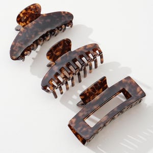 May include: Three tortoiseshell hair claw clips in varying shapes. The clips are brown with dark brown marbling. One clip is curved, one has a series of teeth, and the third is rectangular with a cutout.