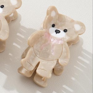 May include: A light brown teddy bear-shaped hair clip with a pink bow. The clip has black eyes and a white muzzle. The bear's body is a marbled, translucent material. The clip is a cute accessory.