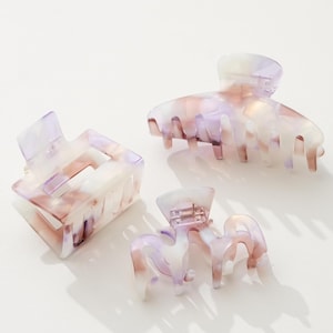 Op de afbeelding: Drie haarclips met marmerpatroon in tinten lavendel, wit en bruin. De clips hebben verschillende maten en vormen, waaronder een rechthoekig en een afgerond ontwerp. De clips zijn gemaakt van een doorschijnend materiaal.