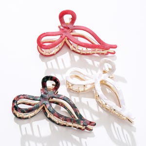 Puede incluir: Tres pinzas para el pelo en rojo, blanco y un estampado floral oscuro. Cada pinza tiene un diseño de lazo con detalles dorados. Las pinzas están hechas de un material brillante y están dispuestas sobre una superficie blanca.