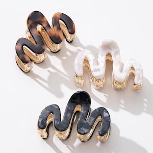 May include: Three wavy-shaped hair claw clips in a variety of colors. One clip is tortoiseshell, another is white with a pearlescent effect, and the third is black and gray. All clips have gold-colored metal accents.