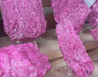 May include: A collection of raw, vibrant magenta-colored mineral specimens displayed on clear acrylic stands. Each piece features a rough, crystalline texture with deep pink hues and jagged edges, arranged across tiered light-colored wooden shelves.