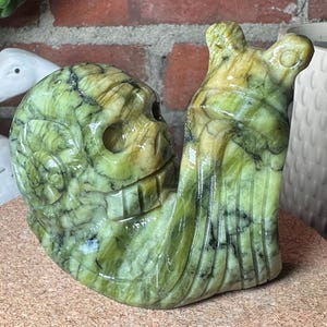 May include: A carved green stone sculpture shaped like a snail with a human skull integrated into its shell. The mineral features variegated shades of olive and lime green with dark veining throughout, showcasing a smooth, polished surface texture.