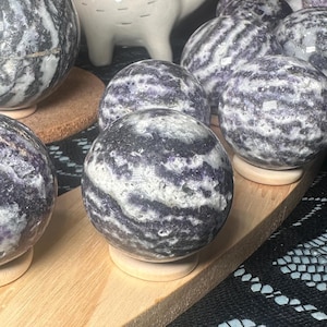 May include: A collection of polished stone spheres in shades of purple, gray, and white. The spheres are displayed on small wooden stands and a wooden tray. The spheres have a marbled appearance and are arranged on a black surface.