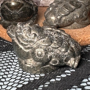 May include: Three carved pyrite frog figurines. The frogs are a dark, metallic gray color with detailed textures. They are arranged on a cork and a black lace surface, with crystals in the background. The frogs are a decorative item.