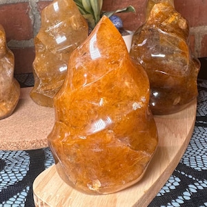 May include: Several polished, amber-colored crystal flame sculptures. The crystals have a smooth, glossy finish and are displayed on a wooden board. The background includes a brick wall and a plant.