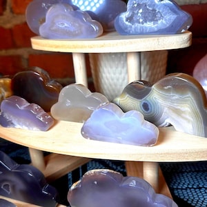 May include: Cloud-shaped agate stones in various shades of purple, gray, and brown are displayed on a two-tiered wooden stand. The stones have unique patterns and textures, with some featuring crystal formations. The background includes a brick wall and a patterned fabric.
