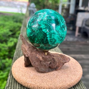 May include: A polished malachite sphere, showcasing vibrant green and dark patterns, is mounted on a natural wood base with a brass stem. The sphere rests on a round, cork-like surface, creating a unique decorative piece.
