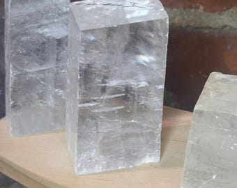 May include: Three clear, rectangular selenite crystal blocks. The crystals are translucent with visible internal striations and a slightly cloudy appearance. They are displayed on a light-colored wooden surface.