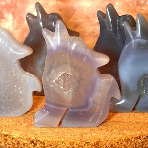 May include: Several carved stone wolf figurines in shades of gray, lavender, and white are displayed. The translucent stone has a layered, banded appearance. The wolf figures are standing on a textured, light brown surface.