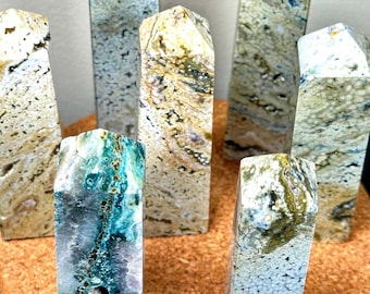 May include: A collection of seven polished stone obelisks. The obelisks display a variety of colors, including beige, green, blue, and brown, with unique patterns and textures. Each obelisk has a rectangular base and a pointed top, showcasing natural stone formations.
