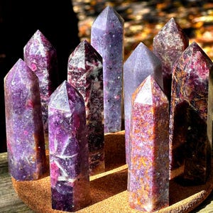 May include: A collection of nine purple crystal points with varying shades and patterns. The crystals are arranged on a round, brown surface, with some crystals appearing translucent. The crystals have pointed tops and are of different heights.
