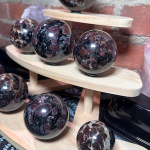 May include: A collection of polished spherical stones featuring deep red and black mottled patterns. These smooth, round mineral spheres are displayed on a tiered light-colored wooden stand, showcasing their reflective surfaces and natural textures.