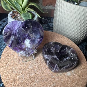 May include: Two heart-shaped amethyst stones with deep purple and white crystalline patterns. One stone is positioned upright on a clear acrylic stand, while the other is a smooth, polished heart-shaped bowl resting flat on a circular cork surface.