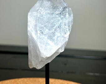 May include: A clear, crystalline quartz crystal, mounted on a black stand. The quartz has a rough, natural texture and is translucent, allowing light to pass through. The crystal is a light, icy color and is displayed against a neutral background.