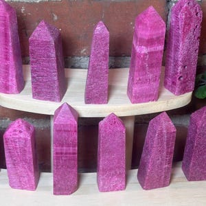 May include: A collection of pink-purple crystal obelisks with textured surfaces and pointed tops. The crystals are displayed on a light-colored wooden shelf and a lower surface. The crystals vary slightly in shape and size, with a natural, raw appearance.