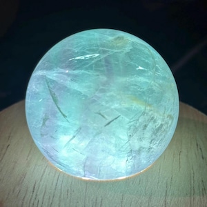 May include: A translucent, spherical crystal ball with a mix of blue and green hues. The crystal has internal lines and inclusions, and it is illuminated from below, creating a glowing effect. The sphere is resting on a wooden surface.
