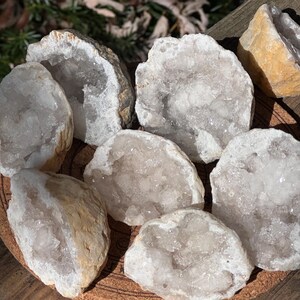 May include: A collection of seven white crystal geodes with a variety of sizes and shapes. The geodes are arranged on a wooden surface.