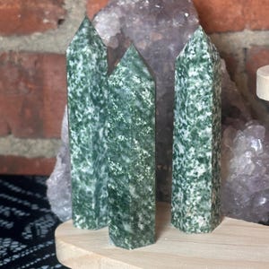 May include: Four green and white speckled crystal obelisks. The obelisks are standing upright on a wooden base. The crystals have a pointed top and a rectangular shape. The background includes a brick wall and a purple crystal cluster.