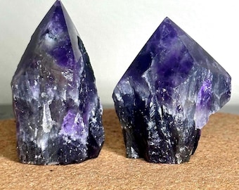 May include: Two raw amethyst crystal points. The crystals are a deep purple color with lighter purple and white inclusions. The rough-cut bases transition to smooth, pointed tips. The crystals are displayed on a light brown surface.