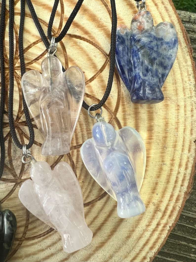 Może przedstawiać: Cztery wiszące ozdoby w kształcie anioła na czarnych sznurkach. Ozdoby wykonane są z czystego kwarcu, r&oacute;żowego kwarcu, niebieskiego agatu i mlecznego niebieskiego kryształu. Ozdoby są prezentowane na drewnianej powierzchni.