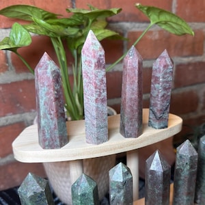 May include: A collection of polished crystal obelisks in varying sizes. The crystals display a mix of red and green hues, with a natural, textured appearance. They are arranged on a light-colored wooden display stand, with a potted plant in the background.