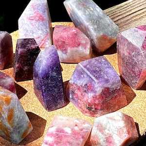 May include: A collection of polished gemstones in shades of purple, pink, and white. The crystals have a variety of shapes, including rectangular and hexagonal forms. The gemstones are displayed on a light brown surface.